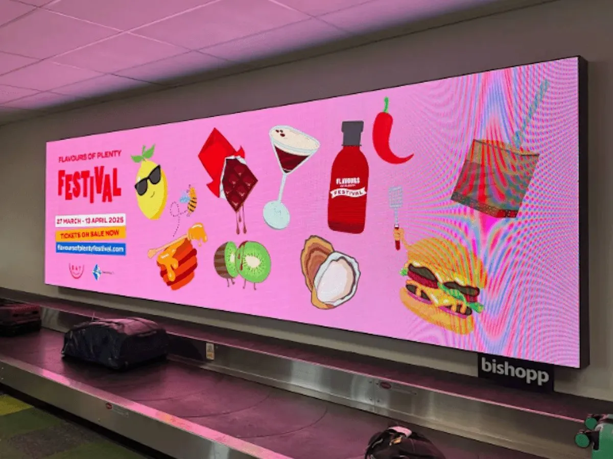 Flavours of Plenty Festival artwork on a digital screen above an airport baggage carousel